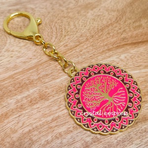 Puede incluir: Llavero dorado con un charm redondo mandala rosa y marrón con un diseño de árbol de la vida dorado.