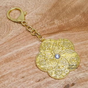 Puede incluir: Llavero dorado con un dije en forma de flor que presenta detalles intrincados y un cristal transparente en el centro. El dije tiene el texto "crystal empress" en la parte inferior.