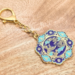 以下が含まれることがあります： 青、ターコイズ、金色のドラゴンとフェニックスのデザインが施された装飾的なチャームが付いた、金色のキーホルダー。 チャームはチェーンと留め金に取り付けられており、木製の背景に配置されています。