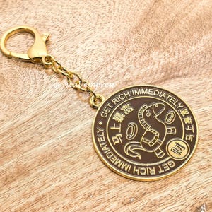 以下が含まれることがあります： 金色のキーホルダー。丸いチャームが付いており、茶色の中心に馬のイラストと「GET RICH IMMEDIATELY」の英語と漢字が書かれています。木製の表面に置かれています。