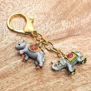 May include: A gold-coloured keyring with two charms: a rhino and an elephant. The rhino and elephant are grey with red, green, and gold accents. The keyring is on a wooden surface.