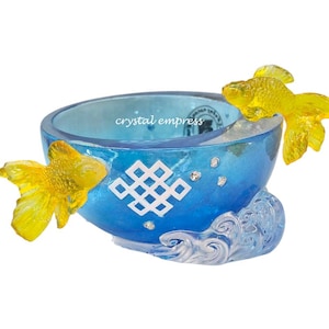 Könnte beinhalten: Eine dekorative blaue Schale mit zwei gelben Goldfischfiguren. Die Schale hat ein weißes geometrisches Design und den Text "crystal empress". Der Sockel hat ein wellenförmiges Design.