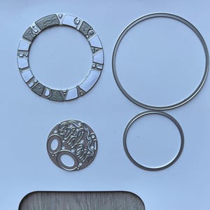 May include: A collection of silver metal die cuts for crafting. Includes a circular frame with the word "Birthday" segmented around the edge, two plain circles, and a small circle with the word "Birthday" in a script font.