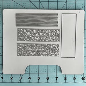 May include: A white die-cut template with three rectangular textured designs: wood grain, pebble, and leopard print patterns. A rectangular frame is also included. The template is on a blue grid surface.