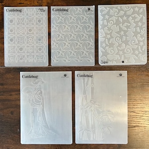 May include: Five Cuttlebug embossing folders with various designs. The designs include a floral pattern, a star pattern, a pattern of stars and swirls, a figure in traditional clothing, and bamboo stalks.