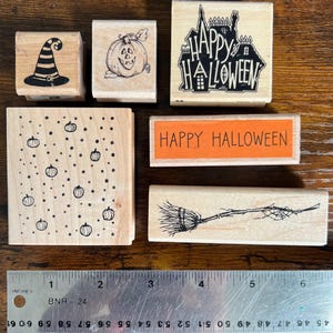 May include: A collection of wooden Halloween-themed stamps. Designs include a witch's hat, jack-o'-lantern, haunted house, pumpkins, the text "Happy Halloween", and a broom. A metal ruler with inch measurements is at the bottom.