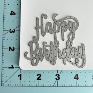 May include: A gray metal die cut with the words "Happy Birthday" in a cursive font. The letters are dotted with small holes. The die cut is on a white card, with a ruler in inches and centimeters in the background.