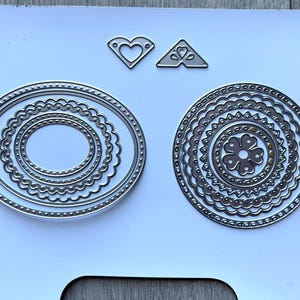 May include: A collection of silver metal die cuts in various shapes. Includes oval and circular frames with decorative edges, and two smaller shapes with heart and triangle designs. Ideal for paper crafting and scrapbooking projects.