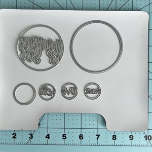 May include: A set of metal die cuts on a white card. The die cuts include a circle with the words "Thinking of You", a plain circle, and three smaller circles with text. The card is on a blue and green grid.