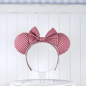 May include: A headband featuring two round ears covered in a red and white gingham fabric. A large, matching red and white gingham bow is centered between the ears. The edges of the ears and the headband are decorated with small white pearls.