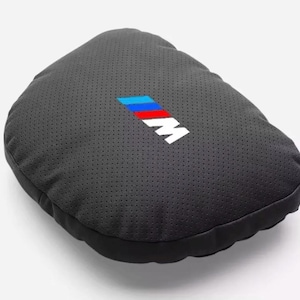 May include: A black, perforated cushion cover with the BMW M logo in blue, red, and white. The cover is a dark gray color and has a rounded shape. The BMW M logo is centered on the top of the cover.