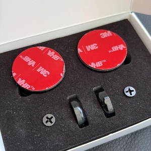 May include: A set of black, metallic car accessories in a white box. The set includes two round, red adhesive pads with "3M VHB" branding, two black rings, and two silver screws. The box is open, revealing the contents.