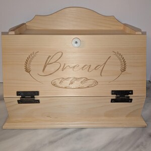May include: A light-colored wooden bread box with a curved top and a hinged door. The word "Bread" is etched on the front, along with wheat stalks and a loaf of bread. The box has a small white knob and black hinges.