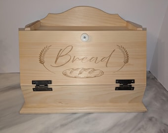 May include: A light-colored wooden bread box with a curved top and a hinged door. The word "Bread" is etched on the front, along with wheat stalks and a loaf of bread. The box has a small white knob and black hinges.
