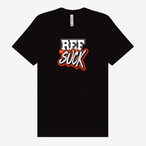 May include: Black t-shirt with the text "REF SUCK" in a white, red, and black design. The word "REF" is in a block letter style, and "SUCK" is in a cursive style with a red outline.