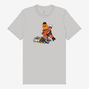 Può includere: Una maglietta grigio chiaro con un'illustrazione a cartoni animati di una mascotte di hockey in arancione, nero e bianco, che sembra attaccare un giocatore di hockey in uniforme nera e gialla. La maglietta è realizzata in un materiale morbido.