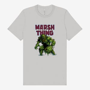 Könnte beinhalten: Hellgraues T-Shirt mit einer Grafik eines grünen Monsters und dem weinroten Text "MARSH THING". Das Monster hat einen Bart und hält einen grünen Gegenstand. Das Shirt hat einen Rundhalsausschnitt.