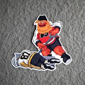 Op de afbeelding: Een gestanste sticker met een pluizig oranje wezen met een zwarte muts en een rood-wit ijshockeytenue, die een ijshockeystick vasthoudt boven een gevallen ijshockeyspeler in een wit shirt met het nummer 87 op de rug en een zwarte broek.