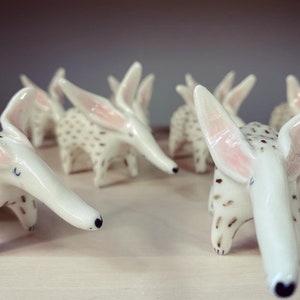 May include: A collection of white ceramic aardvark figurines. Each figurine has long ears with pink interiors, a long snout, and brown spotted markings. The aardvarks are arranged on a light-colored surface.