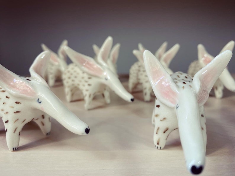 May include: A collection of white ceramic aardvark figurines. Each figurine has long ears with pink interiors, a long snout, and brown spotted markings. The aardvarks are arranged on a light-colored surface.