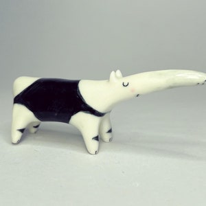 May include: A small ceramic anteater figurine with a glossy white finish, featuring a prominent black patch across its midsection, black-tipped feet, and simple hand-painted facial details including closed eyes and a small pink cheek blush.