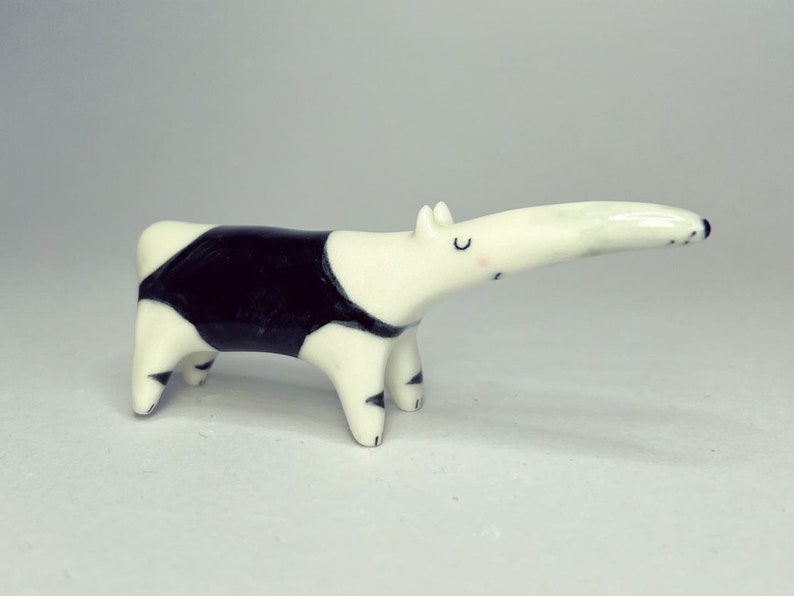 May include: A small ceramic anteater figurine with a glossy white finish, featuring a prominent black patch across its midsection, black-tipped feet, and simple hand-painted facial details including closed eyes and a small pink cheek blush.