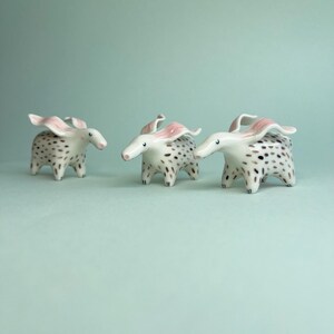 May include: Three ceramic animal figurines with white bodies, black spots, and pink ears. The figurines have long snouts and are posed on a light blue surface. The whimsical creatures are decorative and artistic.