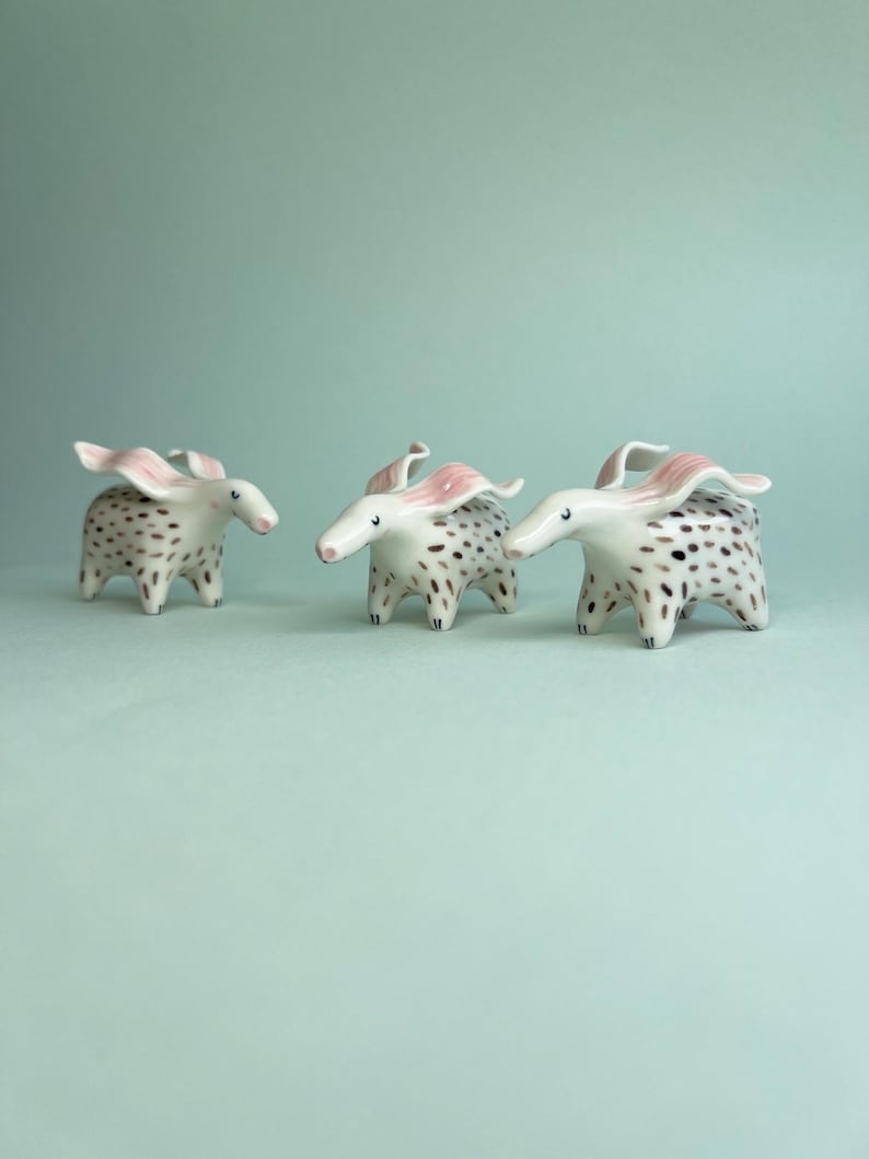 May include: Three ceramic animal figurines with white bodies, black spots, and pink ears. The figurines have long snouts and are posed on a light blue surface. The whimsical creatures are decorative and artistic.