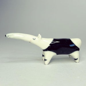 May include: A small ceramic figurine of a Malayan tapir with a glossy white and black finish. The animal is depicted in a standing position with a long snout, small ears, and closed eyes, featuring a distinct black patch across its midsection.