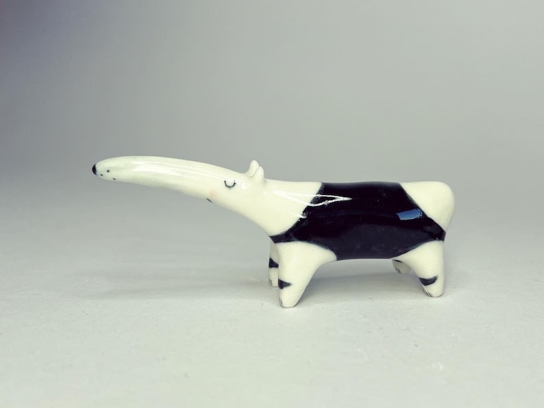 May include: A small ceramic figurine of a Malayan tapir with a glossy white and black finish. The animal is depicted in a standing position with a long snout, small ears, and closed eyes, featuring a distinct black patch across its midsection.