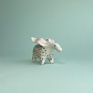May include: A ceramic figurine of a stylized animal with a white body, brown spots, and pink ears. The figurine has a small, rounded body with four short legs and a small, pink nose. The background is a solid, light blue-green color.
