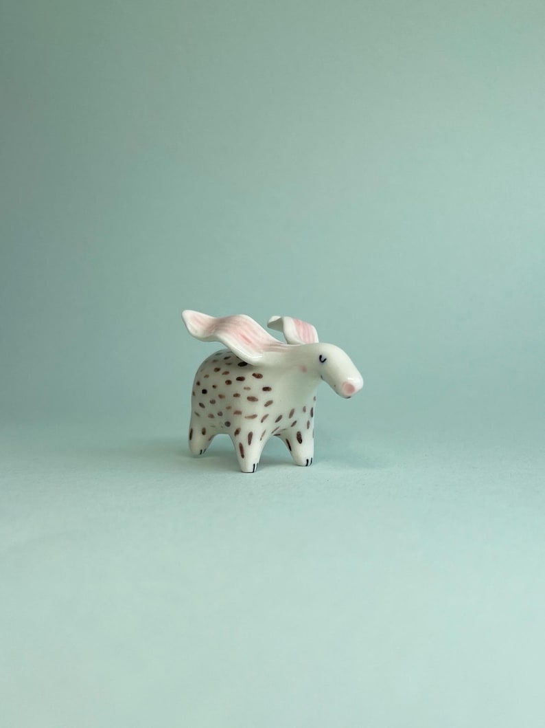 May include: A ceramic figurine of a stylized animal with a white body, brown spots, and pink ears. The figurine has a small, rounded body with four short legs and a small, pink nose. The background is a solid, light blue-green color.