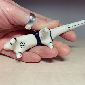 May include: A small, cream-colored ceramic animal figurine with a long tail and black markings. The underside features a pattern of small black dots on the belly and the number 217 painted in black near the hind legs. The surface is smooth and glossy.