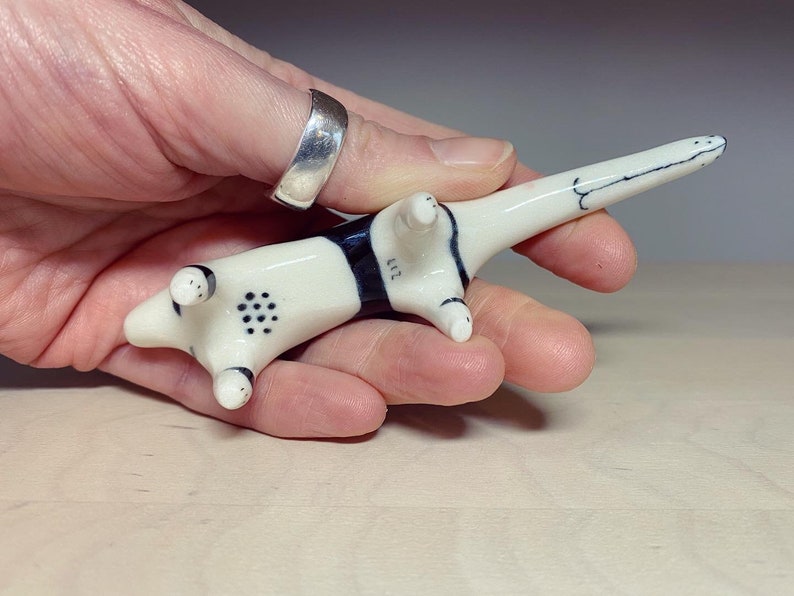 May include: A small, cream-colored ceramic animal figurine with a long tail and black markings. The underside features a pattern of small black dots on the belly and the number 217 painted in black near the hind legs. The surface is smooth and glossy.