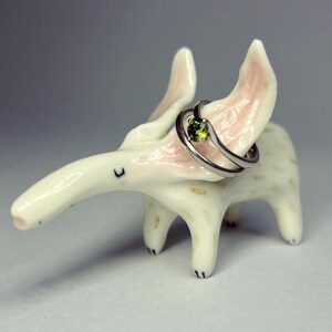 May include: A whimsical, off-white ceramic anteater ring holder with pink-tinged ears and a long snout. A silver ring with a green gemstone is displayed on the anteater's back. The ring holder has black painted details.
