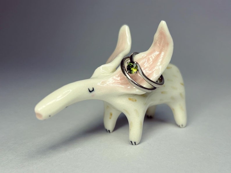 May include: A whimsical, off-white ceramic anteater ring holder with pink-tinged ears and a long snout. A silver ring with a green gemstone is displayed on the anteater's back. The ring holder has black painted details.