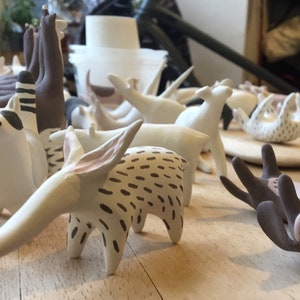 May include: A collection of handmade ceramic animal figurines, including a raccoon, deer, and other stylized creatures. The figurines are primarily white with brown and black accents, arranged on a wooden surface.