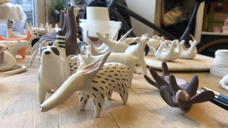 May include: A collection of handmade ceramic animal figurines, including a raccoon, deer, and other stylized creatures. The figurines are primarily white with brown and black accents, arranged on a wooden surface.