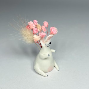 Puede incluir: Figura de conejo de cerámica blanca con vientre rosa y detalles negros. El conejo sostiene un ramo de flores rosas y un acento beige plumoso. Artículo decorativo.