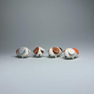 Puede incluir: Cuatro pequeñas figuras de cerámica de cobayas. Cada figura es blanca con manchas grises, marrones y naranjas. Las cobayas están en fila, con sus pequeñas patas visibles.