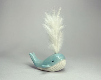 May include: A light blue ceramic whale figurine with a white feathery plume on top. The whale has a simple, smiling face.