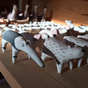 May include: A collection of small, hand-painted ceramic animal figurines arranged in rows. The foreground features an anteater with black and white markings alongside a spotted animal with pinkish-brown spots on a cream-colored base.