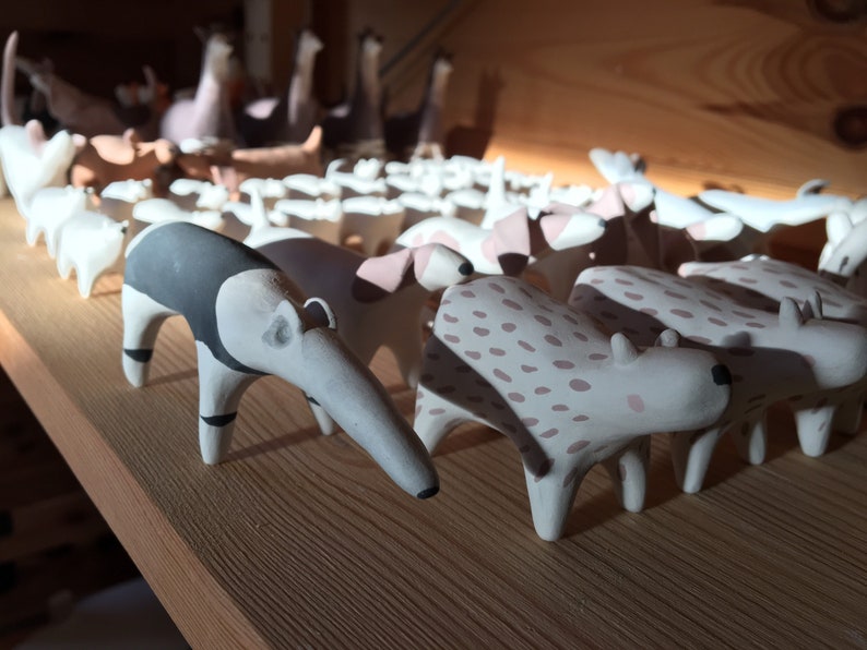 May include: A collection of small, hand-painted ceramic animal figurines arranged in rows. The foreground features an anteater with black and white markings alongside a spotted animal with pinkish-brown spots on a cream-colored base.