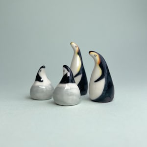 Puede incluir: Cuatro figuras de pingüinos de cerámica. Los pingüinos tienen cuerpos negros y blancos con detalles amarillos en sus picos. Las figuras varían en tamaño y están dispuestas sobre un fondo azul claro. Los pingüinos son un artículo decorativo.