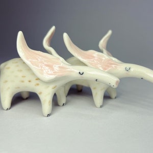 May include: Two whimsical ceramic creatures with long, curved snouts and large, pink-lined ears. The bodies are off-white with brown spots, and the legs are short and rounded. The creatures have small, black eyes and are posed together.