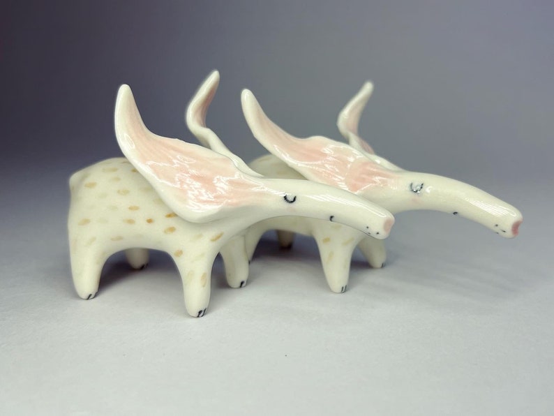 May include: Two whimsical ceramic creatures with long, curved snouts and large, pink-lined ears. The bodies are off-white with brown spots, and the legs are short and rounded. The creatures have small, black eyes and are posed together.