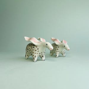 May include: Two ceramic animal figurines. The figurines are white with brown spots and pink ears. The animals have small black feet and are standing on a light green surface.