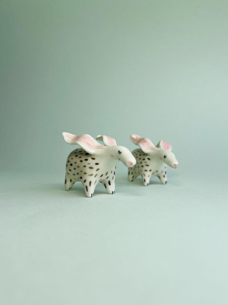 May include: Two ceramic animal figurines. The figurines are white with brown spots and pink ears. The animals have small black feet and are standing on a light green surface.