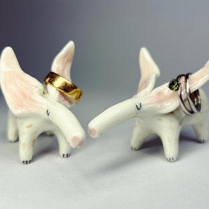 May include: Two whimsical ceramic elephant ring holders. One holds a gold ring, the other holds silver rings with a green gemstone. The elephants are white with pink accents on their ears. The ring holders are decorative and functional.