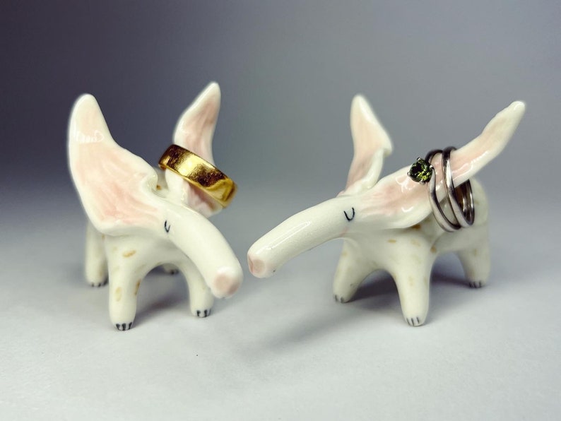 May include: Two whimsical ceramic elephant ring holders. One holds a gold ring, the other holds silver rings with a green gemstone. The elephants are white with pink accents on their ears. The ring holders are decorative and functional.